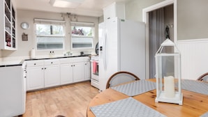 Une cuisine avec des meubles blancs, un réfrigérateur, une table à manger avec des sets de table et une fenêtre avec des stores.