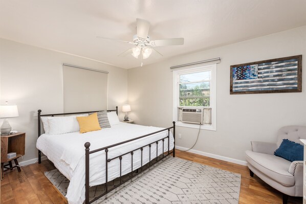 Une chambre à coucher comprenant un lit, des tables de chevet, un ventilateur de plafond, une fenêtre avec un climatiseur et une chaise.