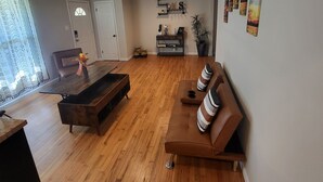 A spacious living area with a wooden floor, a brown sofa, a coffee table, and a dining table. There is a door with a window, a wall-mounted shelf with decorative items, and a potted plant. - HOT TUB SUNFLOWER OASIS HOME (Sherman)