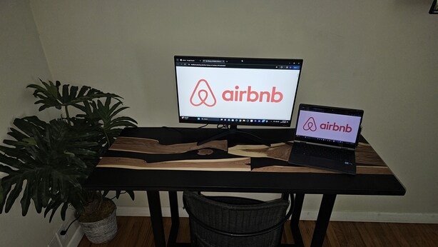 A wooden desk with two monitors displaying the Airbnb logo, a potted plant, and a wicker basket. - HOT TUB SUNFLOWER OASIS HOME (Sherman)