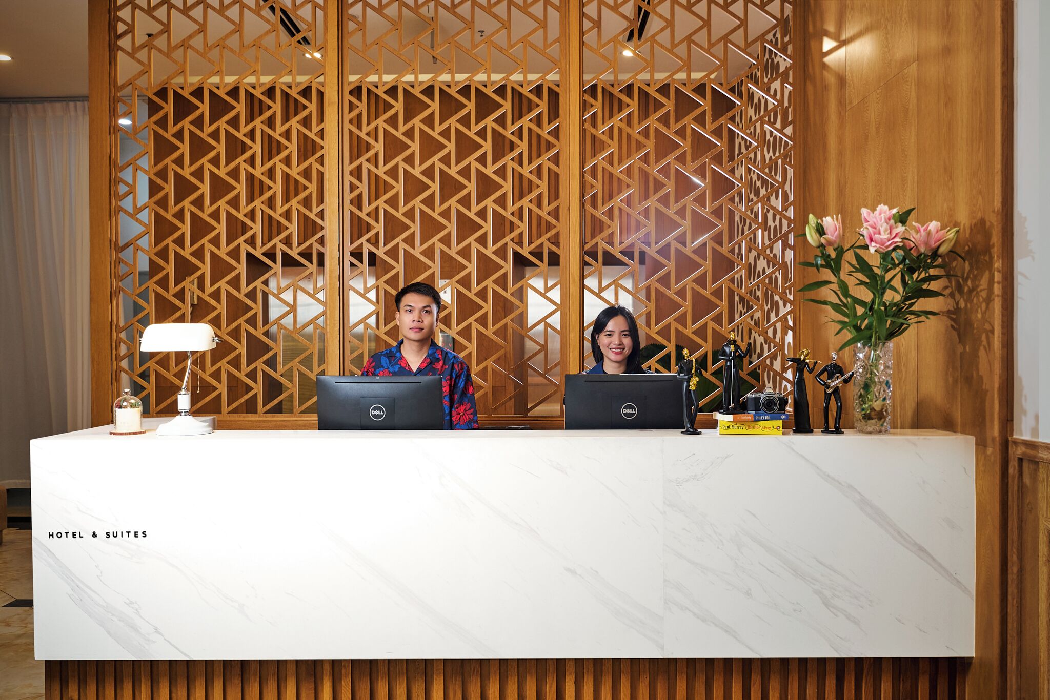 Un comptoir d’accueil d’hôtel avec deux employés, un comptoir en marbre et un paravent décoratif en bois.