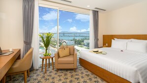 Hotelkamer met een groot bed, een bureau met een laptop, een stoel, een klein tafeltje met een plant, en een uitzicht op de stad en zee door grote ramen.