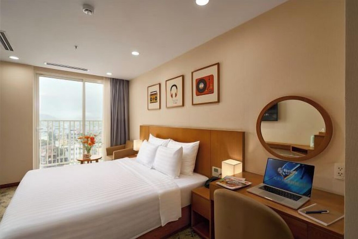 Une chambre d’hôtel avec un grand lit, un bureau avec un ordinateur portable, un miroir rond et des tableaux encadrés accrochés au mur.