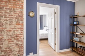 Un pasillo con pared de ladrillo, una puerta azul que da a un dormitorio y una estantería con diversos objetos.