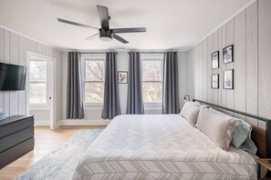 Une chambre à coucher avec un lit, un ventilateur de plafond, une commode, un téléviseur et une fenêtre avec des rideaux.