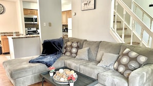 Ein Wohnzimmer mit einem grauen Ecksofa, einem Couchtisch mit Dekoration und einer Küche im Hintergrund.