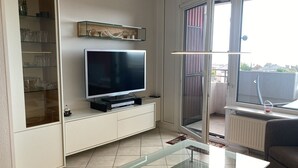 Una sala moderna con un televisor de pantalla plana sobre un mueble blanco, una mesa de centro de vidrio y vistas al exterior a través de un ventanal corredero.
