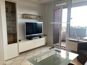 Una sala moderna con un televisor de pantalla plana sobre un mueble blanco, una mesa de centro de vidrio y vistas al exterior a través de un ventanal corredero.