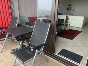 Una sala moderna con puerta de vidrio que da a un balcón, un comedor con mesa y sillas, y una cocina con un armario blanco y una planta.
