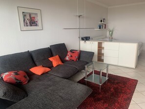A modern living room with a grey sectional sofa, red and black patterned cushions, a glass coffee table, and a red shag rug. - Berolina 510 - 90 degrees east - course 90° east - direct view of the Baltic Sea! (Dahme)