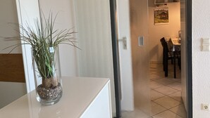 Una habitación con un escritorio blanco, una planta en maceta y una puerta que da a un comedor con mesa y sillas.