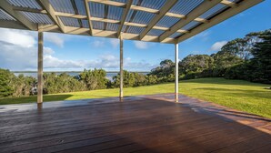 Un espace extérieur abrité, doté d’une terrasse en bois et d’un toit en métal, offrant une vue sur un paysage verdoyant et une étendue d’eau.