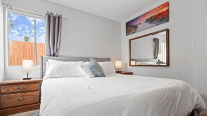 Un dormitorio con una cama grande, mesitas de noche de madera, una ventana con cortinas, un cuadro y un espejo en la pared.