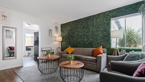 Un salón moderno con una pared verde, dos mesas redondas, un sofá gris y un ventanal grande.