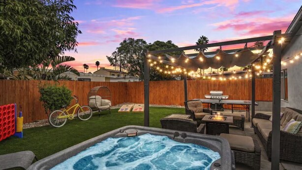 Un patio trasero con un jacuzzi, mobiliario exterior, un cerco de madera y luces colgantes.