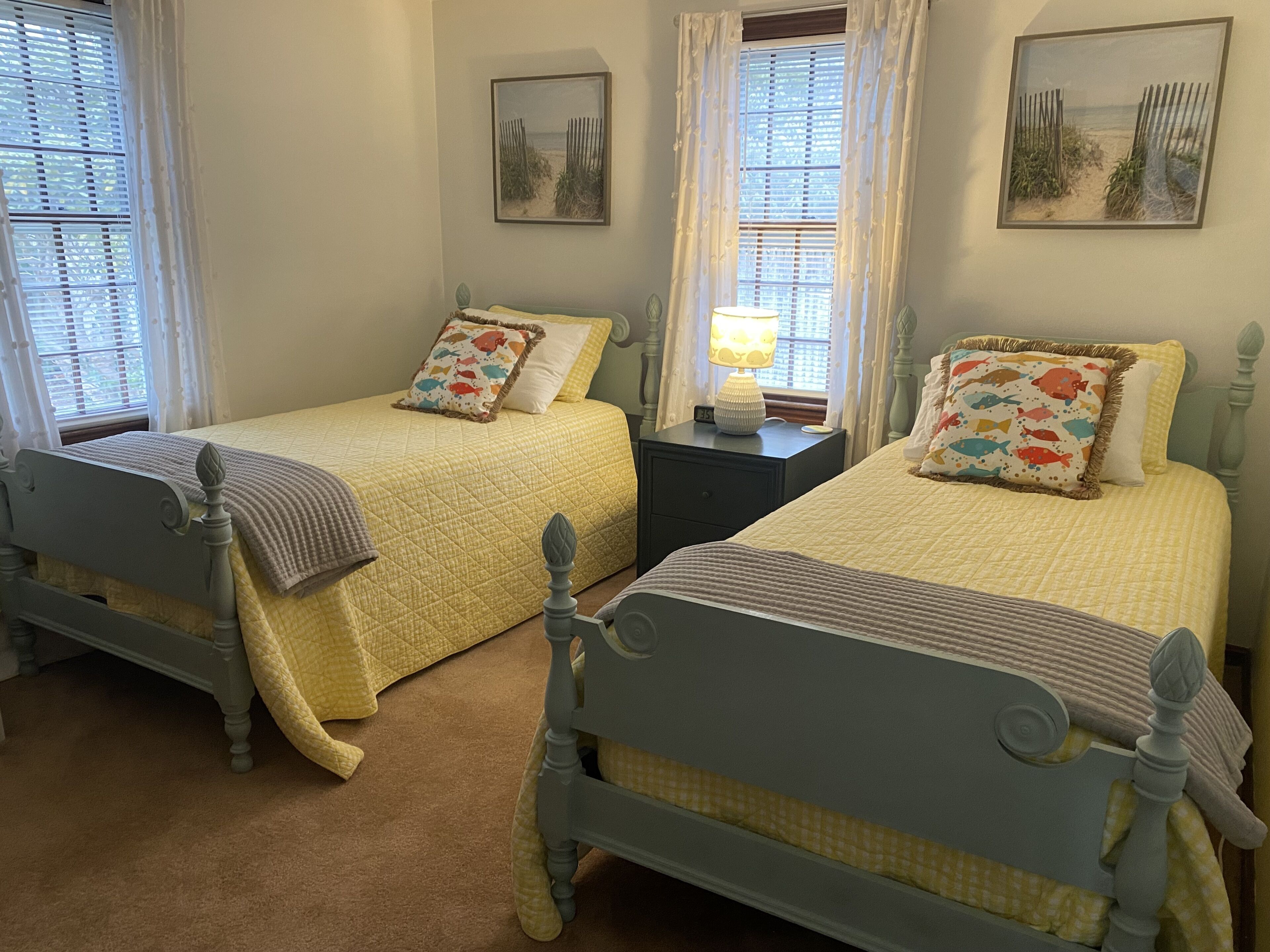 A bedroom with two twin beds, each with a headboard, a bedside table with a lamp, and a window with curtains.