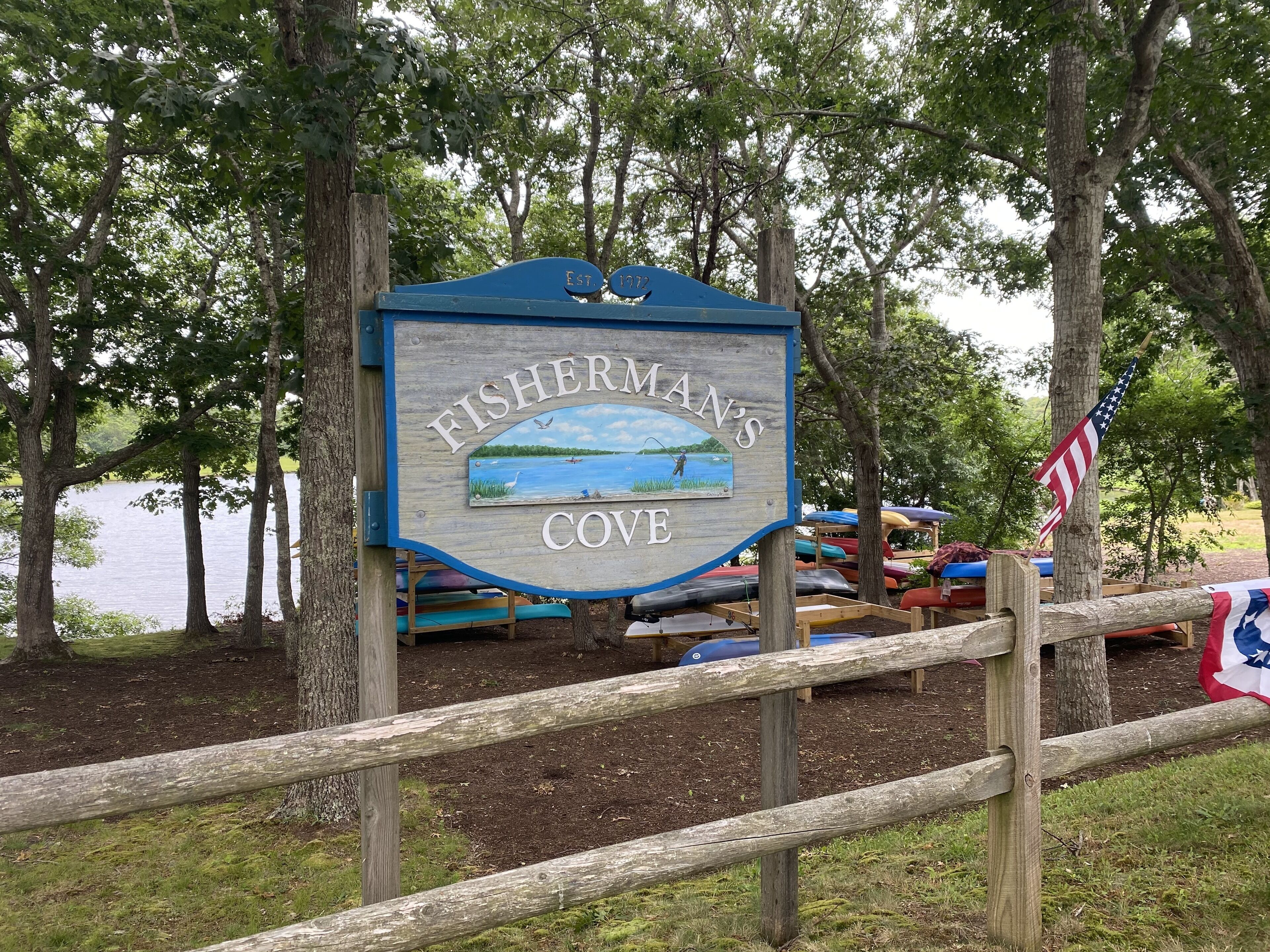 Ein hölzernes Schild mit der Aufschrift "FISHERMAN'S COVE", umgeben von Bäumen und einem Gewässer.