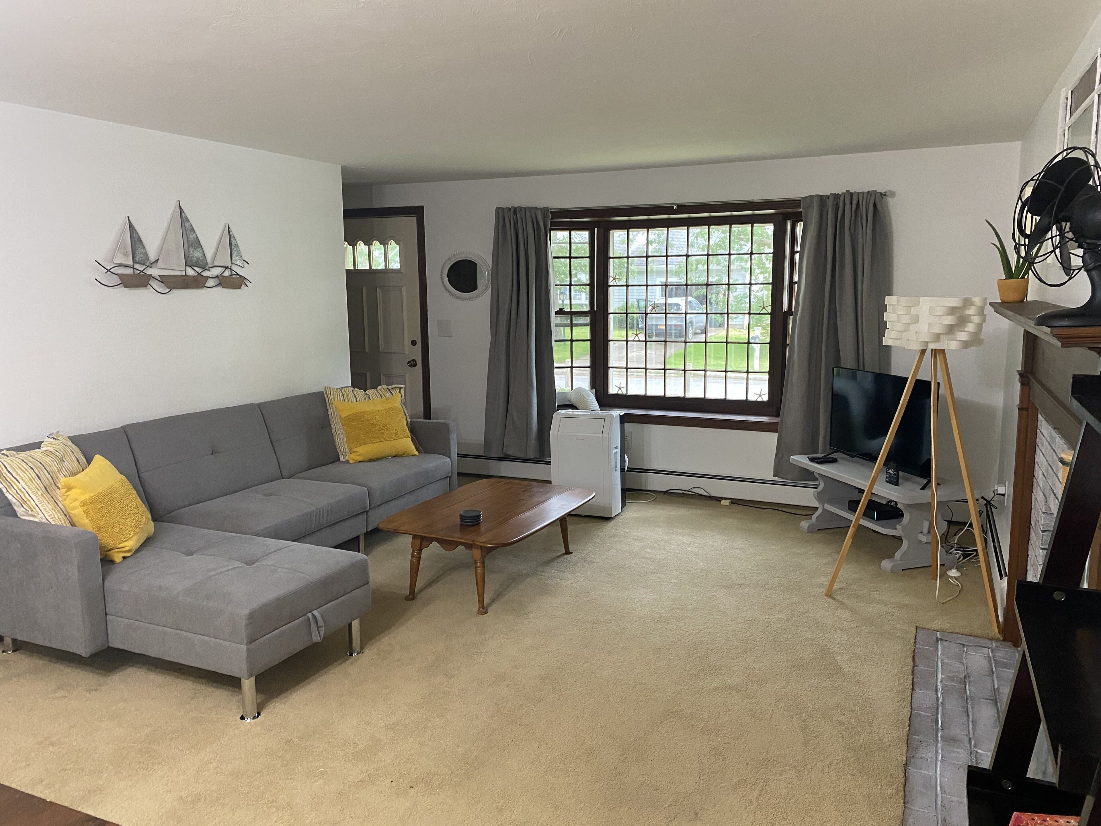 Een woonkamer met een grijze bank, gele kussens, een houten salontafel, een televisie op een meubel en een groot raam met gordijnen.