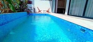 Ein Schwimmbecken mit klarem, blauem Wasser, umgeben von einer gefliesten Kante und einer weißen Wand. In der Nähe des Beckens befinden sich zwei Holzstühle und ein kleiner Tisch.