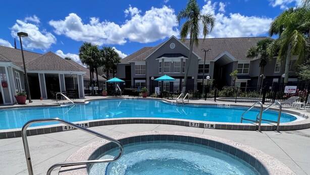 A swimming pool with a hot tub, surrounded by lounge chairs and umbrellas, with a building in the background.