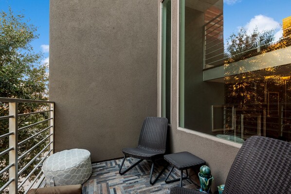 A balcony with a wicker chair, a small ottoman, and a glass table.