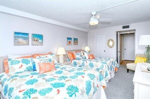 Une chambre avec deux lits, un ventilateur de plafond et des œuvres d’art murales représentant des scènes de plage.