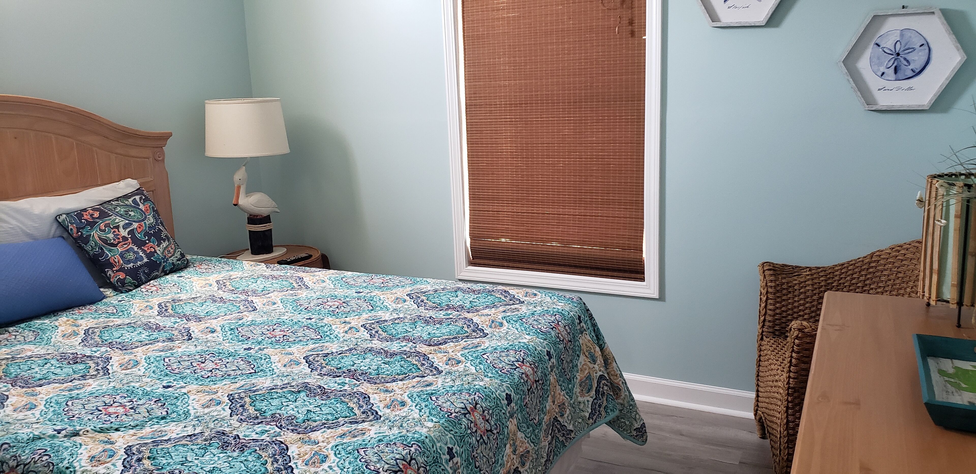 Una camera da letto con una coperta a motivi, una testiera in legno, una lampada da comodino, una finestra con persiane e una sedia in vimini.