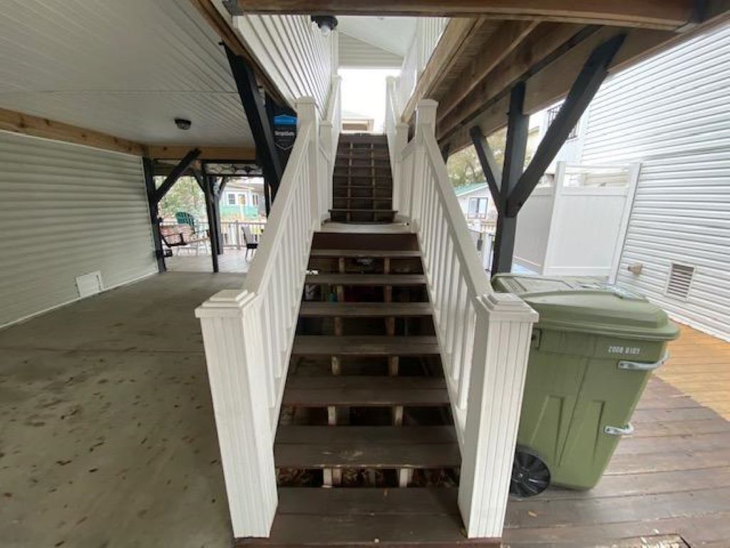Una escalera exterior techada con escalones de madera y barandillas blancas, que conduce a un área cubierta con un bote de basura verde.