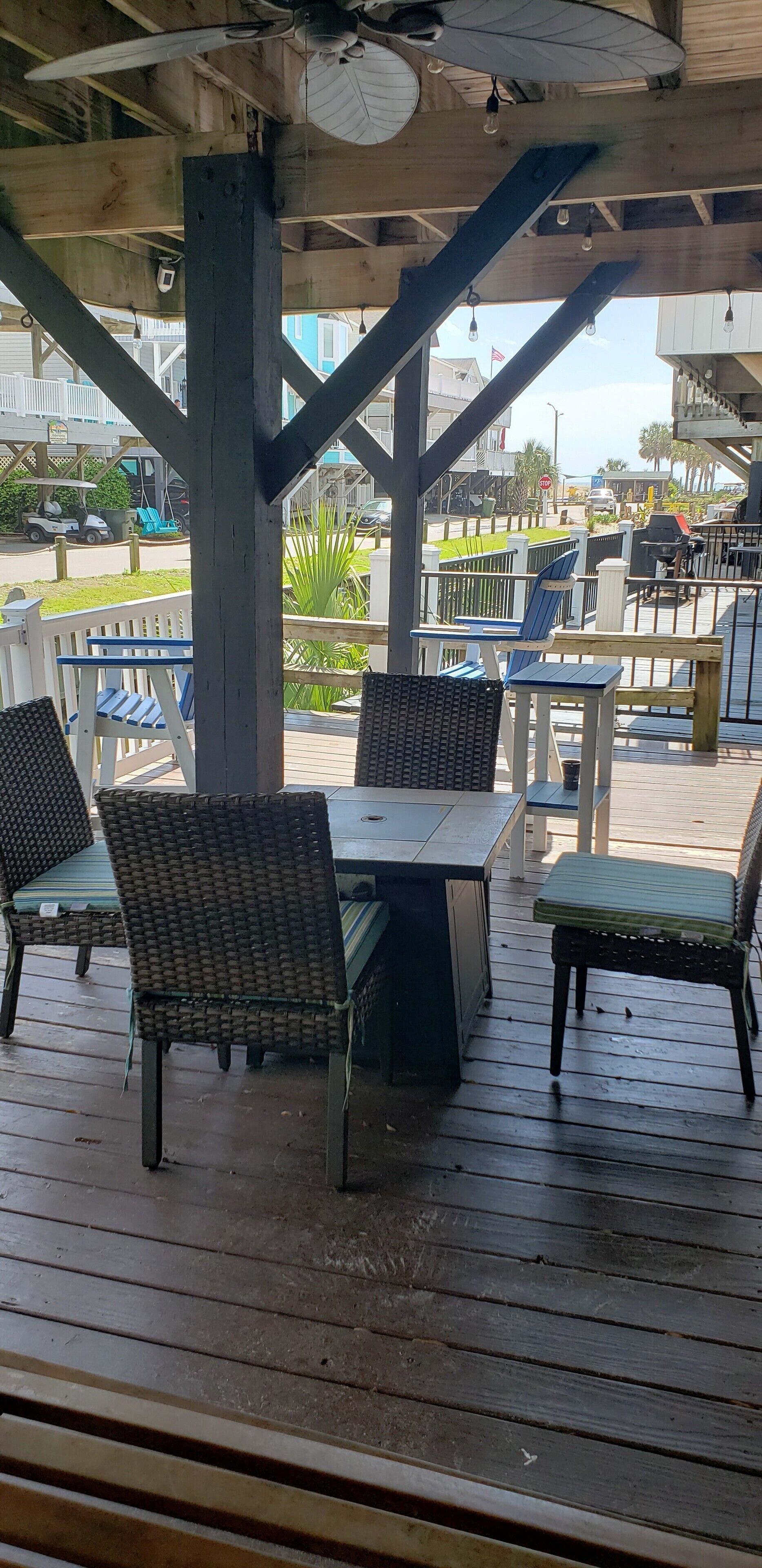 Un espace extérieur aménagé, abrité, avec des chaises et des tables en osier, une terrasse en bois et une vue sur la rue et les bâtiments en arrière-plan.