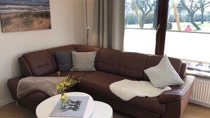 A brown leather sofa with a white throw blanket and two pillows, a small white table with a vase of yellow flowers, and a painting of a beach scene on the wall.