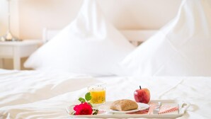 A neatly made breakfast tray with a glass of orange juice, a red apple, a croissant, and a pink rose on a bed with white bedding and pillows. - Apartment No.3 - Seestern - Ferienhof Kluvetasch (Neustadt in Holstein)