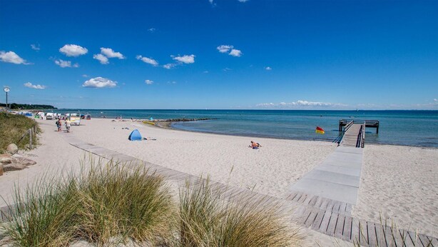 A beach with a wooden boardwalk leading to the water, a tent, and a flag on the sand. - Apartment No.3 - Seestern - Ferienhof Kluvetasch (Neustadt in Holstein)