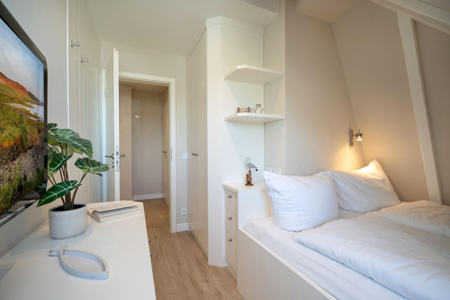 Un dormitorio con una cama, una mesita de noche, un estante en la pared, una puerta que da a otra habitación y una planta en maceta.