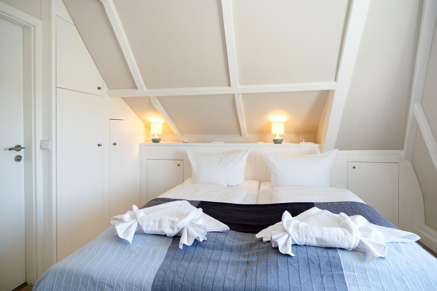 A hotel room with a bed, two bedside lamps, and a door.