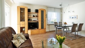 A modern living room with a brown leather sofa, a glass coffee table, a wooden entertainment unit with a flat-screen TV, and a dining area with a glass table and metal chairs. - Risgap - Schmidtpott - Cozy 3-room apartment for 4 persons with terrace (Wenningstedt-Braderup (Sylt))
