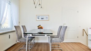A dining area with a glass table, grey upholstered chairs, a fruit bowl, pendant lights, and a framed picture on the wall. - Risgap - Schmidtpott - Cozy 3-room apartment for 4 persons with terrace (Wenningstedt-Braderup (Sylt))