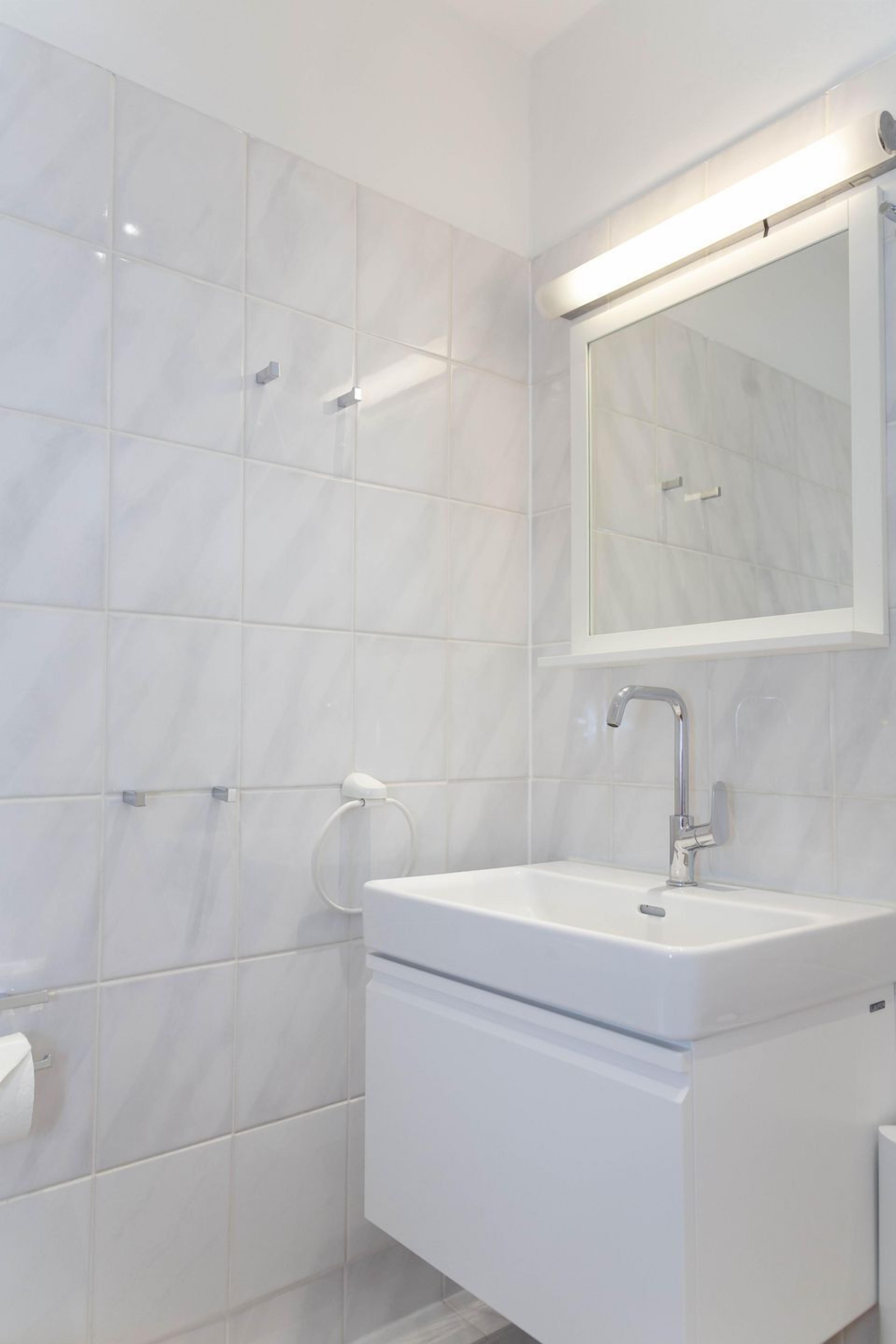 A modern bathroom with white marble tiles, a white sink, and a mirror with integrated lighting.