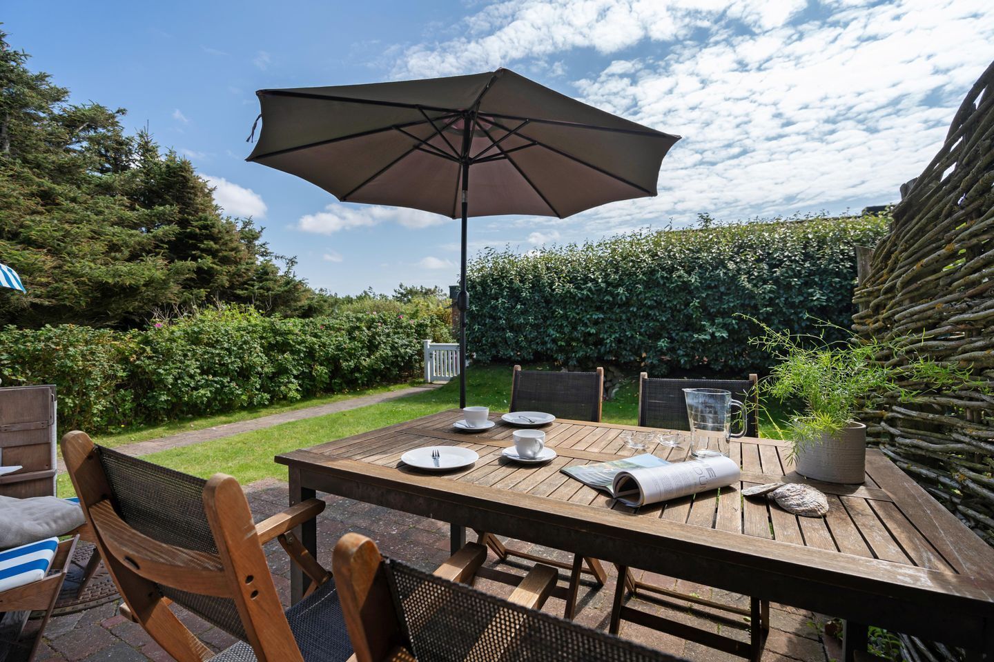 A wooden table set outdoors with a large umbrella, chairs, and a potted plant.
