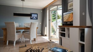 A dining area with a chessboard, chairs, and a table. There is a TV on a shelf and a window with curtains. - Am Sandwall Ostseite - House close to the beach for 4 persons with sauna, terrace, garden, (Sylt)