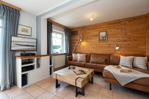 A cozy living room with a wooden wall, a brown leather sofa, a wooden coffee table with chess, and a TV on a white stand.