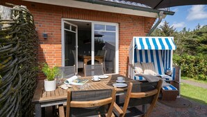 A patio with a wooden table and chairs, a striped beach chair, and a brick wall. - Am Sandwall Ostseite - House close to the beach for 4 persons with sauna, terrace, garden, (Sylt)