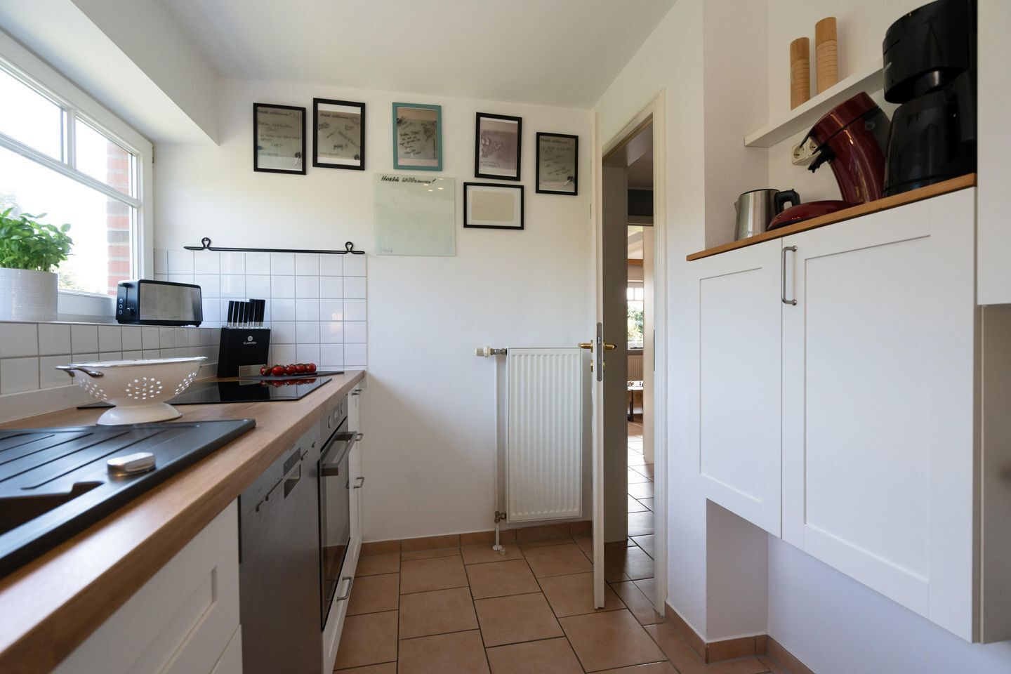 A kitchen with a microwave, toaster, and kettle on the countertop, a white sink, and a window with a plant. There are framed certificates on the wall and a door leading to another room.