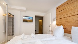 A hotel room with a bed, a wooden headboard, a wall-mounted TV, a bedside table with a lamp, and a closet with hanging clothes. - Am Sandwall Ostseite - House close to the beach for 4 persons with sauna, terrace, garden, (Sylt)