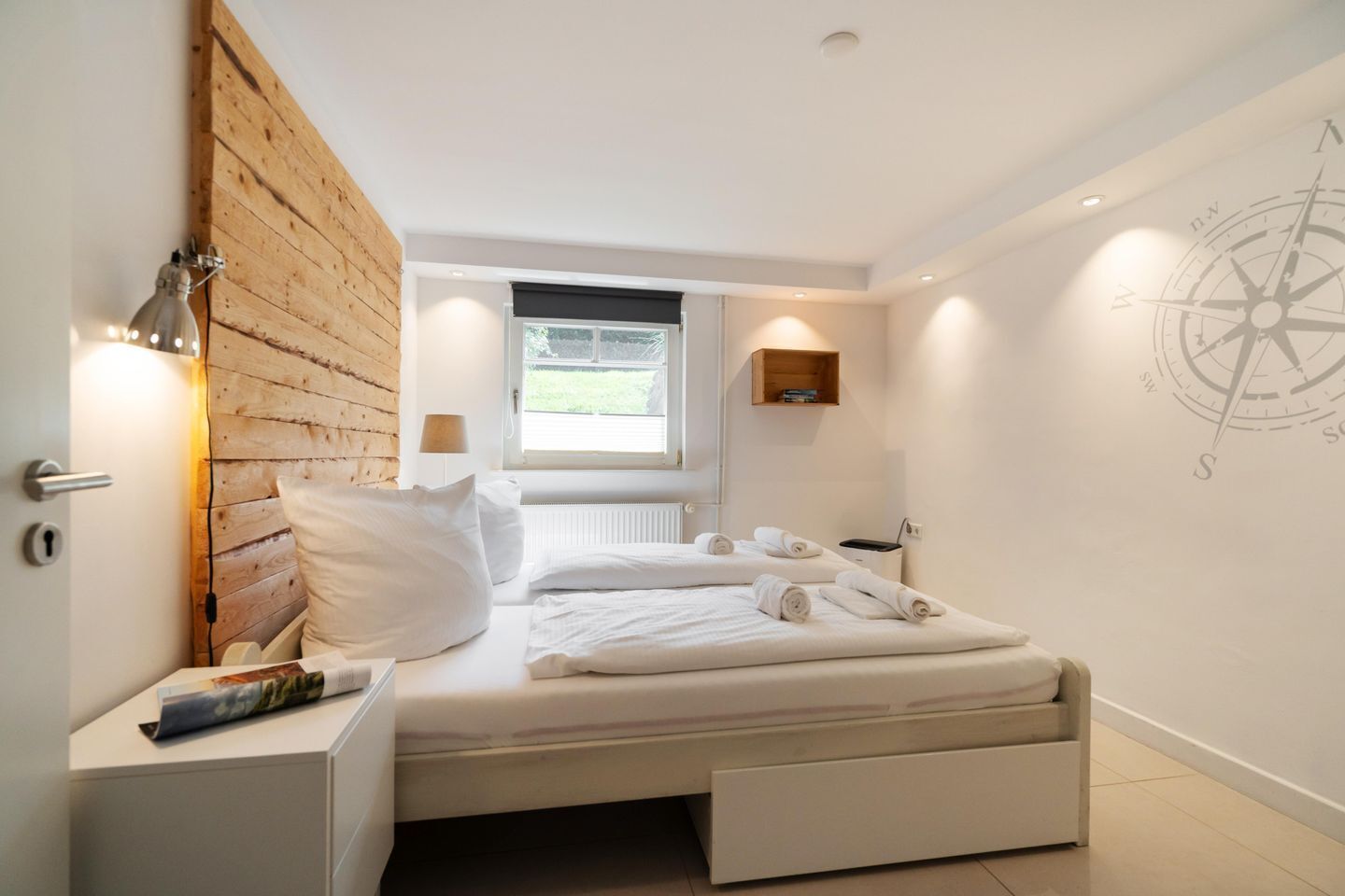 Un dormitorio moderno con cabecera de madera, una ventana con vistas, un adorno de pared con forma de brújula y camas ordenadas con toallas.