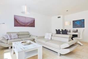 A modern living room with a beige sofa, a white coffee table, and a dining area with a white table and chairs. - Neue Liebe - Modern house for 6 pers. with terrace, parking space, WLAN. (Sylt)