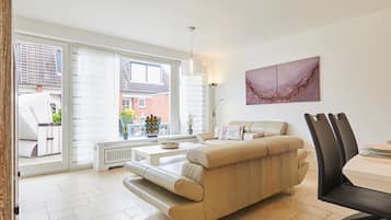 A modern living room with a beige sofa, a dining table with chairs, and a large window offering a view of the outside.