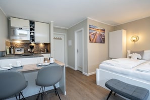 Ein kompaktes Hotelzimmer mit Küchenzeile, einem Bett mit weißer Bettwäsche, einer Bank und einem Sonnenuntergangsbild an der Wand.