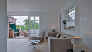 Ein modernes Wohnzimmer mit Sofa, Sessel und Couchtisch. Ein Schiebetürfenster führt zu einem Außenbereich mit einer Terrasse und Blick auf ein Gebäude und Bäume.