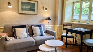 A living room with a grey sofa, two round white side tables, a wooden dining table with chairs, a framed picture on the wall, and a window with blinds. - Kiebitz - Beautiful 2-room apartment with terrace, parking space, WLAN. Pets (Sylt)