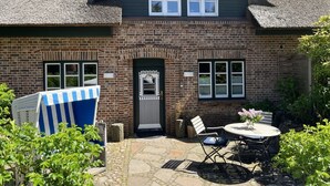 A brick house with a thatched roof, a front door, and a small patio with a table and chairs. - Kiebitz - Beautiful 2-room apartment with terrace, parking space, WLAN. Pets (Sylt)